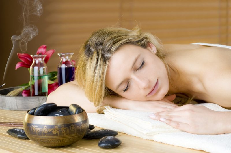 Eine junge Frau entspannt während einer Massage in schönem Ambiente: Räucherduft, Hot Stones und eine Schale sind zu sehen.